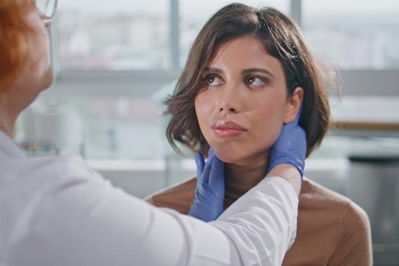 A nurse wearing a white jacket and blue exam gloves and is touching the side of a young woman’s neck