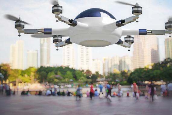 A blue and white drone with six propellers hovers in the foreground, a city plaza filled with people and skyscrapers fills the background 