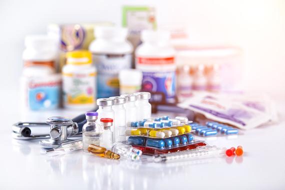 Stacks of packaged capsules, syringes and small bottles with liquid medicine are in front of several blurred bottles of medications