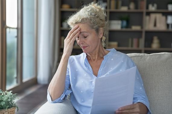 A woman is sitting on a couch and holding documents. Her eyes are closed and she is touching her forehead. She appears to be worried