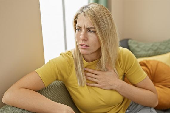 A young woman sits sideways on a couch. She is looking away, frowning and appears worried. Her hand is on her upper chest