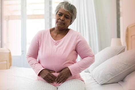 An older woman sits on her bed while grabbing her abdomen with a pained expression on her face