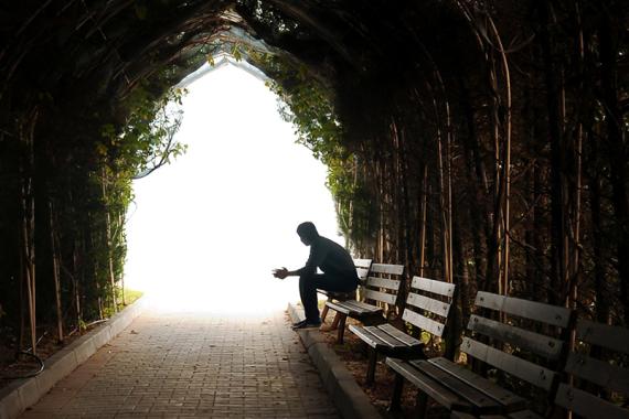 A person sits alone on a bench at the end of a dark stone tunnel. Ivy grows on the tunnel walls. The tunnel opens to a lighted area