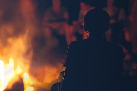 Silhouette of the back of a person sitting in front of a campfire at night. More people are visible on the other side of the campfire