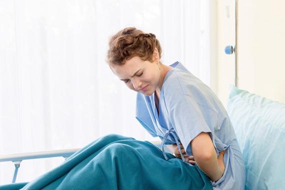 A young woman sitting in a hospital bed. She is leaning forward with her hands over her stomach. She is distressed and in pain