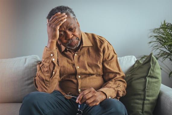 A middle-aged man is sitting on a couch with both hands covering his stomach. He looks like he is in pain.
