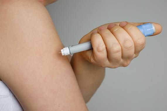 A closeup of a person’s hand and upper arm. The person is holding an injection device and giving themselves an injection in the upper arm
