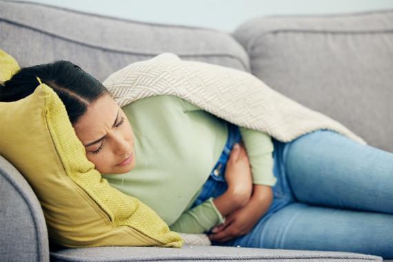 A woman wearing jeans and a light green sweater holds her abdomen in discomfort as she lies on a gray couch, with a yellow throw pillow and beige throw blanket