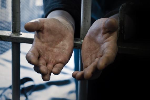 Two open hands extend through metal prison bars. The palms face upward, showing lined, calloused and weathered skin