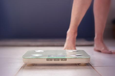 A digital scale sits on the floor in the foreground, in the background a pair of legs indicates a person is about to step onto the scale