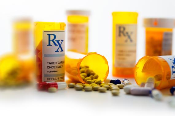 Eight prescription bottles are on a table. The three bottles are in front and two are tipped over with pills spilling onto the table