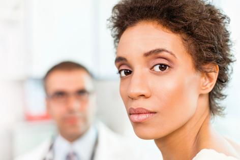 A young woman is in front of a white background, looking forward. A blurred image of man with a stethoscope is behind her
