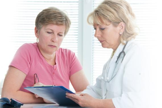 A nurse in a white lab coat is talking to a woman wearing a pink shirt. They both are looking at an open file folder
