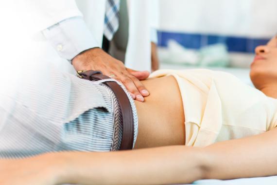 A doctor examines a young female patient lying on an exam table in a doctor’s office. The doctor is touching the patient’s abdomen