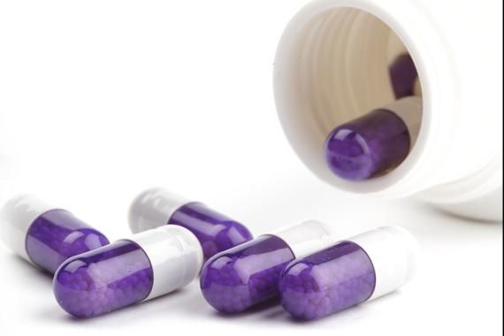 Several purple and white capsules have spilled out of a white bottle onto a white table. Another capsule is about to fall out of the bottle