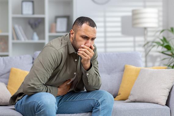 A young man is sitting on a couch. One hand is on his stomach and the other is covering his mouth. He looks nauseated