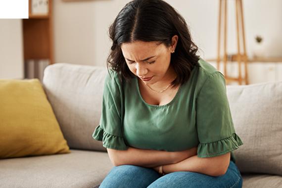 A woman doubles over in pain, crossing her arms in front of her abdomen as she sits on a couch