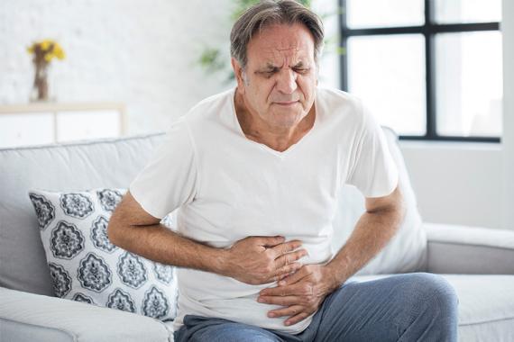 A man wearing a white shirt sits on a couch and holds his abdomen with both his hands and a pained expression on his face