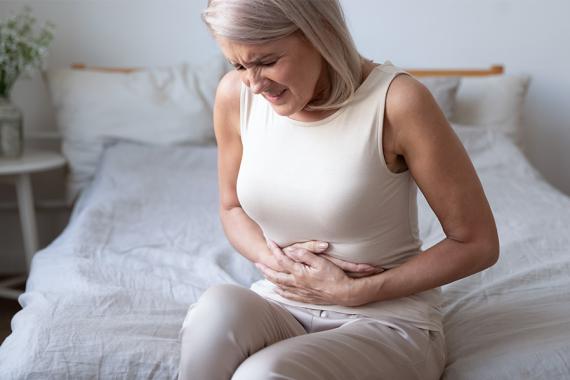 An older woman sits on her bed while grabbing her abdomen with a pained expression on her face