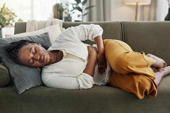 A woman lies on a green couch with throw pillows while holding her abdomen in pain