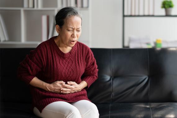 A middle-aged woman sits on a black couch. She has both hands on her stomach and has an expression of pain on her face