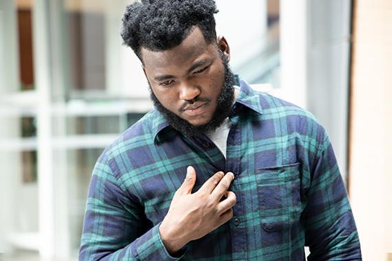A young man is standing with his right hand placed on the middle of his chest. He is looking down with an expression of concern