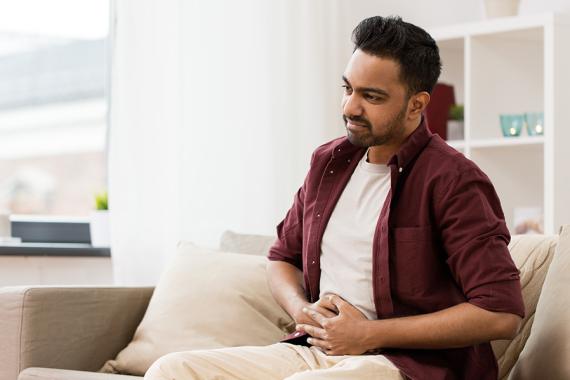 A man sits on a couch and holds his abdomen with a pained expression on his face