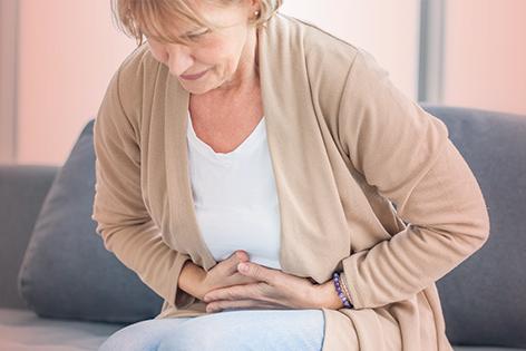 A middle-aged woman is sitting on a couch. She is leaning forward and has both hands on her stomach as if she is having intense stomach pain