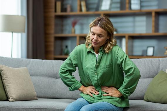 A young woman is sitting on a couch and has both hands on her stomach. She has her eyes closed and looks very uncomfortable